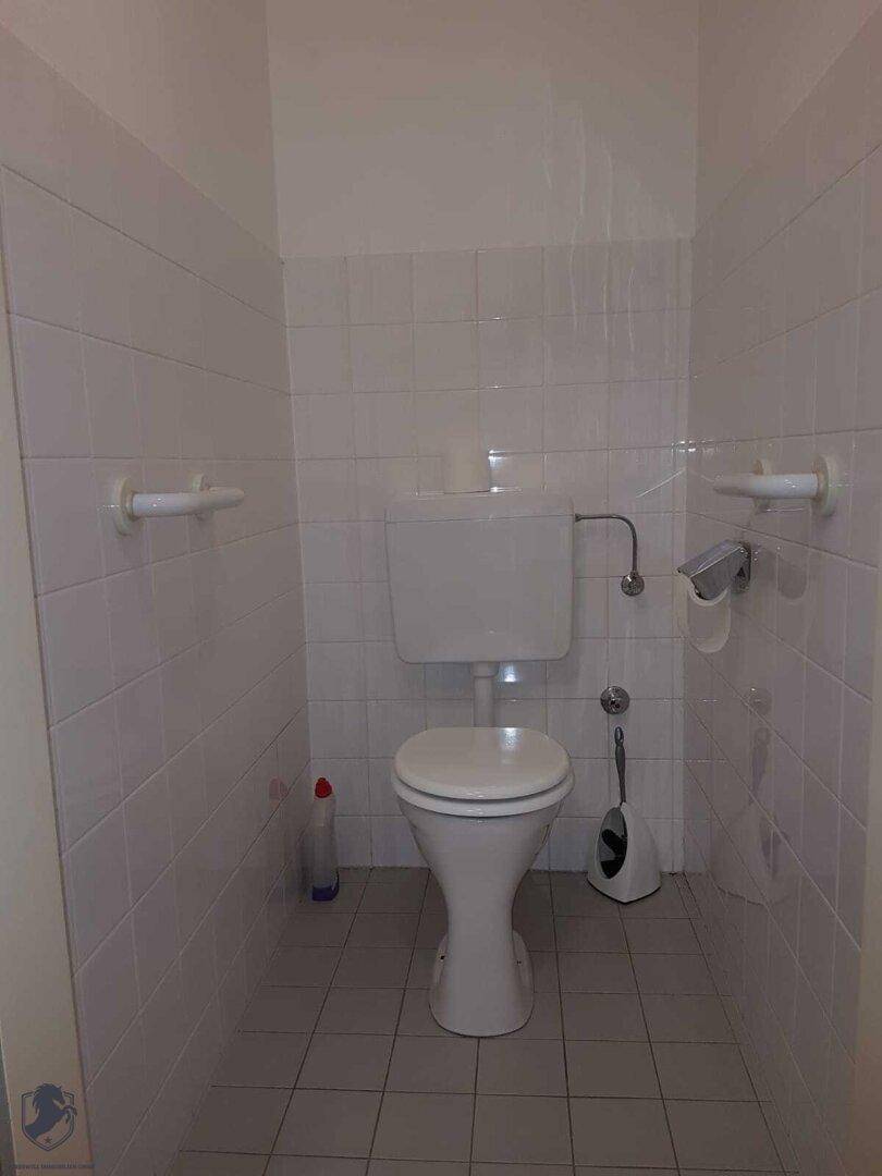 Separates WC mit wandmontierter Toilette, weißen Fliesen und Haltegriffen für Barrierefreiheit.
