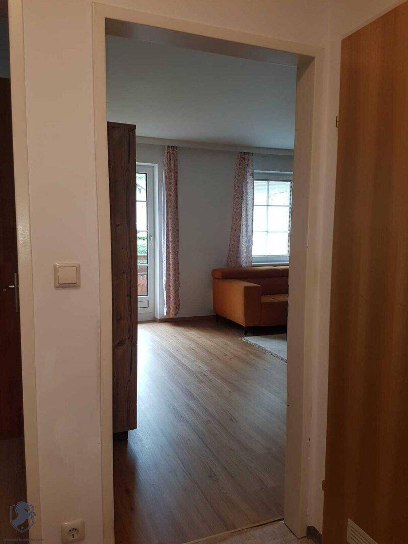 Blick durch einen Türrahmen in ein helles Wohnzimmer mit Holzboden, Sofa und großen Fenstern.