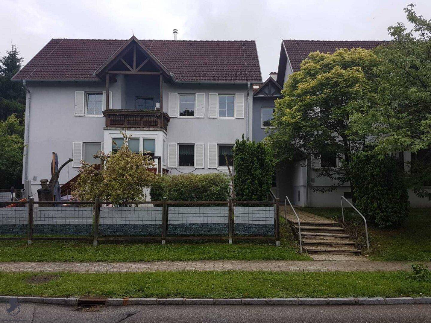 Mehrstöckiges Gebäude mit Satteldach, Balkonen und weißen Fensterläden, umgeben von Bäumen.