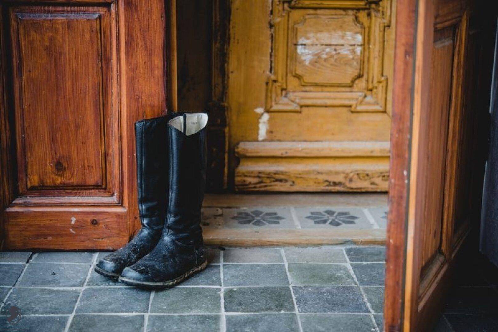Schwarze Stiefel stehen auf einem gefliesten Boden vor rustikalen Holztüren im Eingangsbereich.