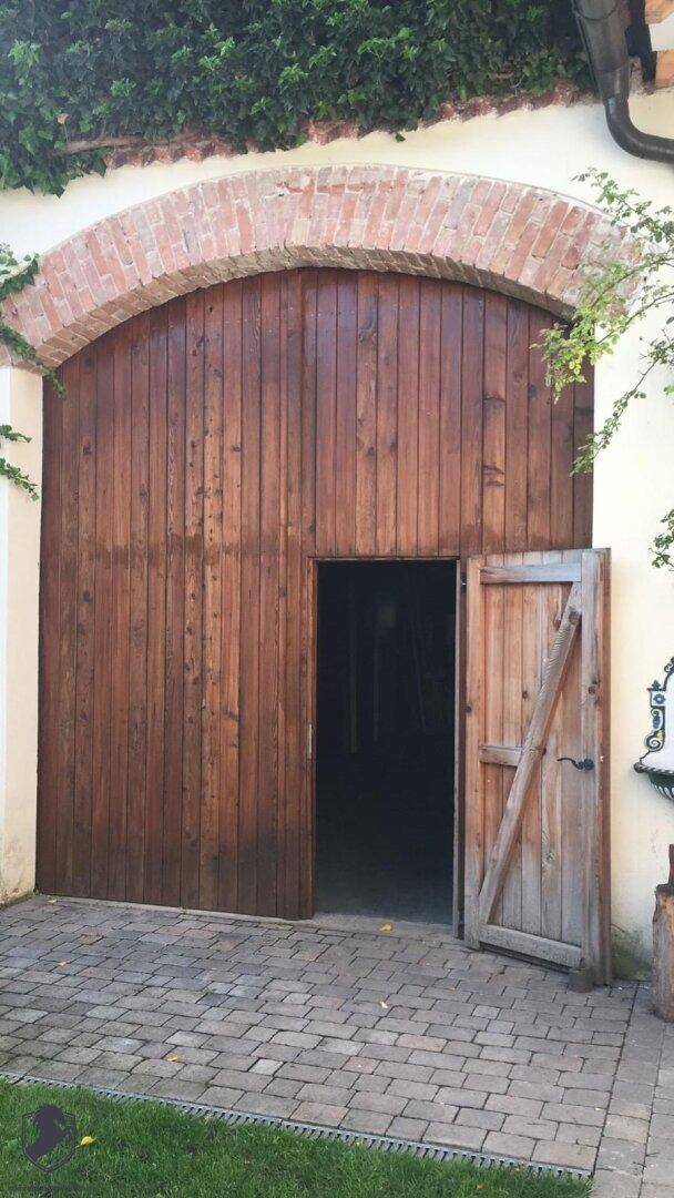 Große, gewölbte Holztür mit einer kleineren geöffneten Zugangstür in einer Ziegelmauer.