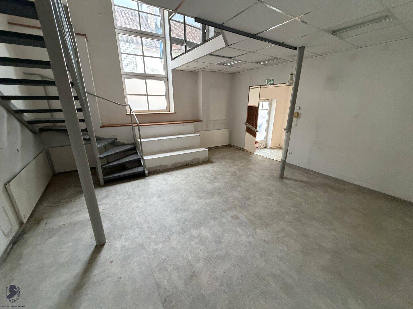 Heller Gewerberaum im Erdgeschoss mit großen Fenstern, Treppe zur oberen Ebene und Betonboden.