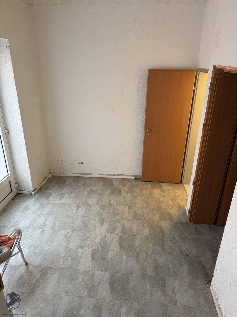Leerer Raum mit hellgrauem Fliesenboden, weißen Wänden und einer Holztür, mit Fensterzugang.