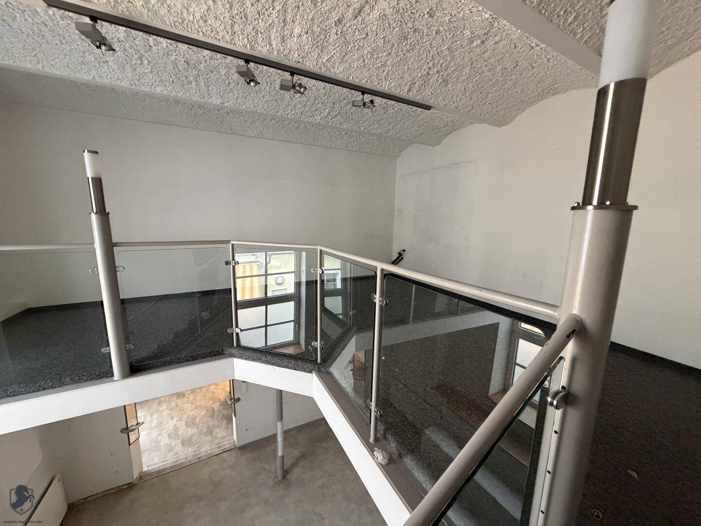 Obere Galerieebene mit modernem Glasgeländer, strukturiertem Deckenputz und Schienenbeleuchtung.
