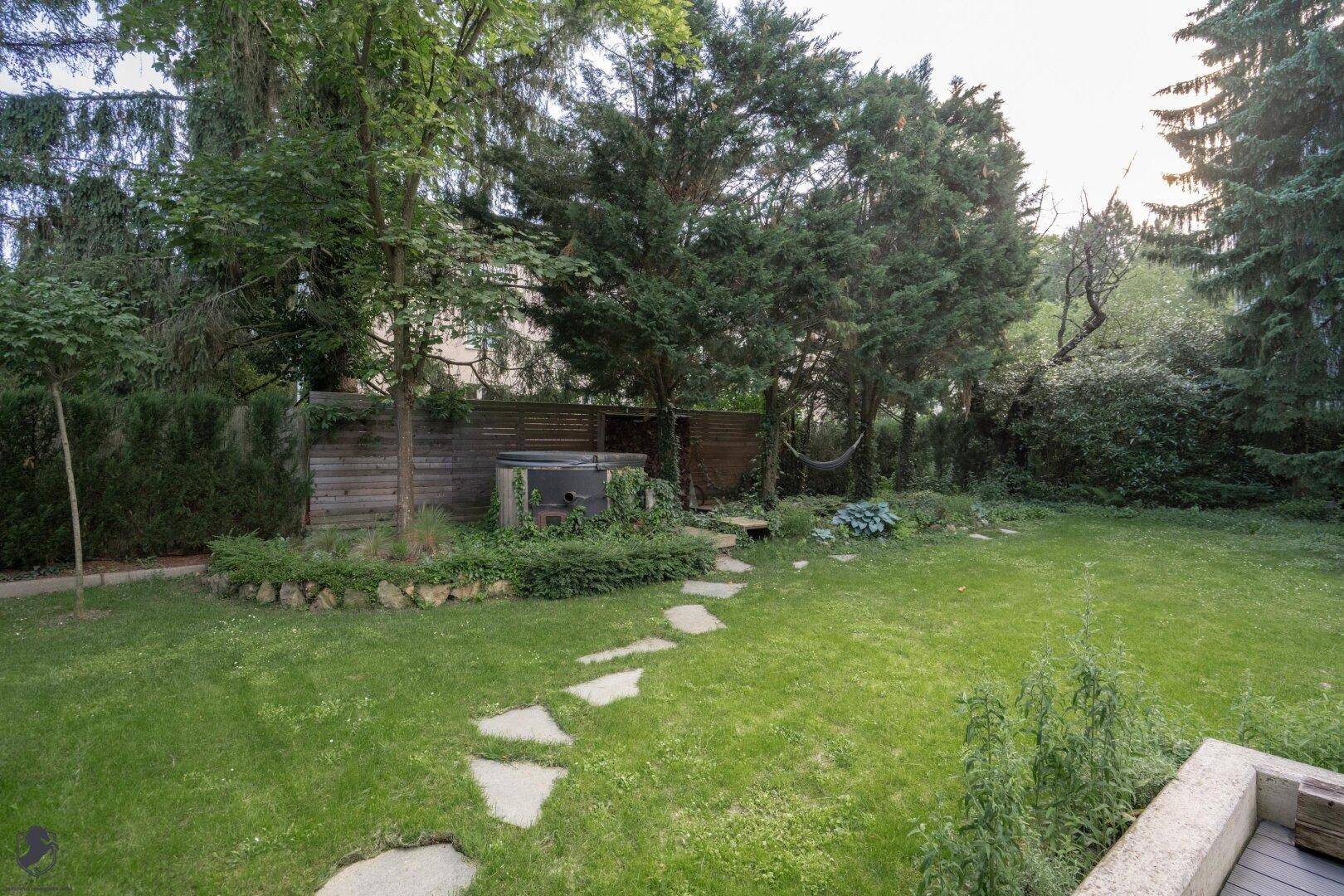Großer, grüner Garten mit Bäumen, Trittsteinen und einem Whirlpool im hinteren Bereich.