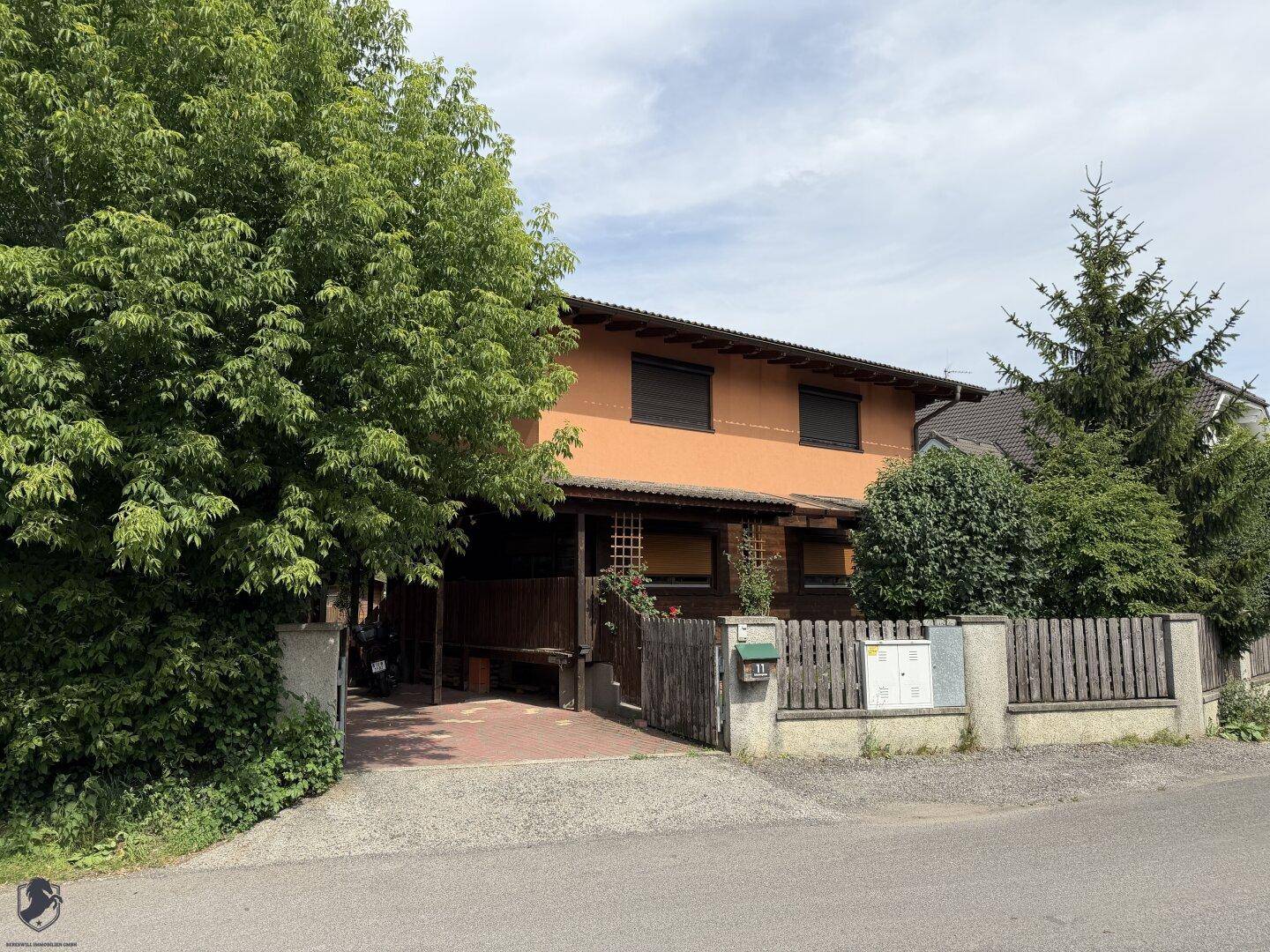 Vorderansicht des Hauses mit orangefarbener Fassade, umgeben von Bäumen und einem Holzzaun.