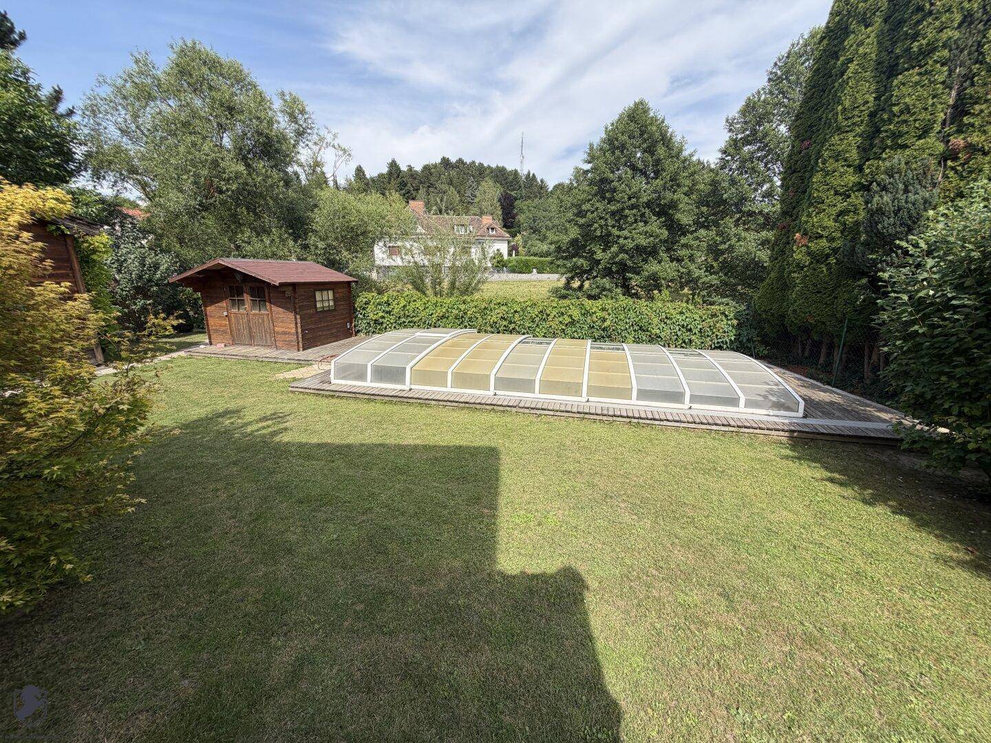 Großer Gartenbereich mit Rasenfläche, überdachtem Pool und einem kleinen Holzschuppen im Hintergrund.
