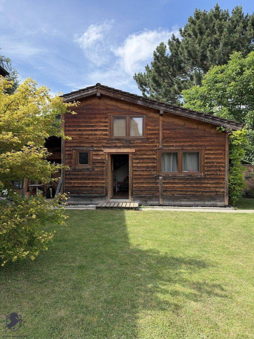 Rustikales Holzhaus im Garten mit grüner Rasenfläche und hohen Bäumen im Hintergrund.