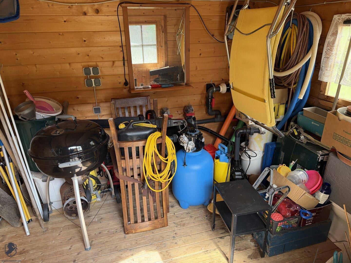 Innenansicht eines Holzschuppens mit Gartengeräten, Grill und verschiedenen Lagergegenständen.