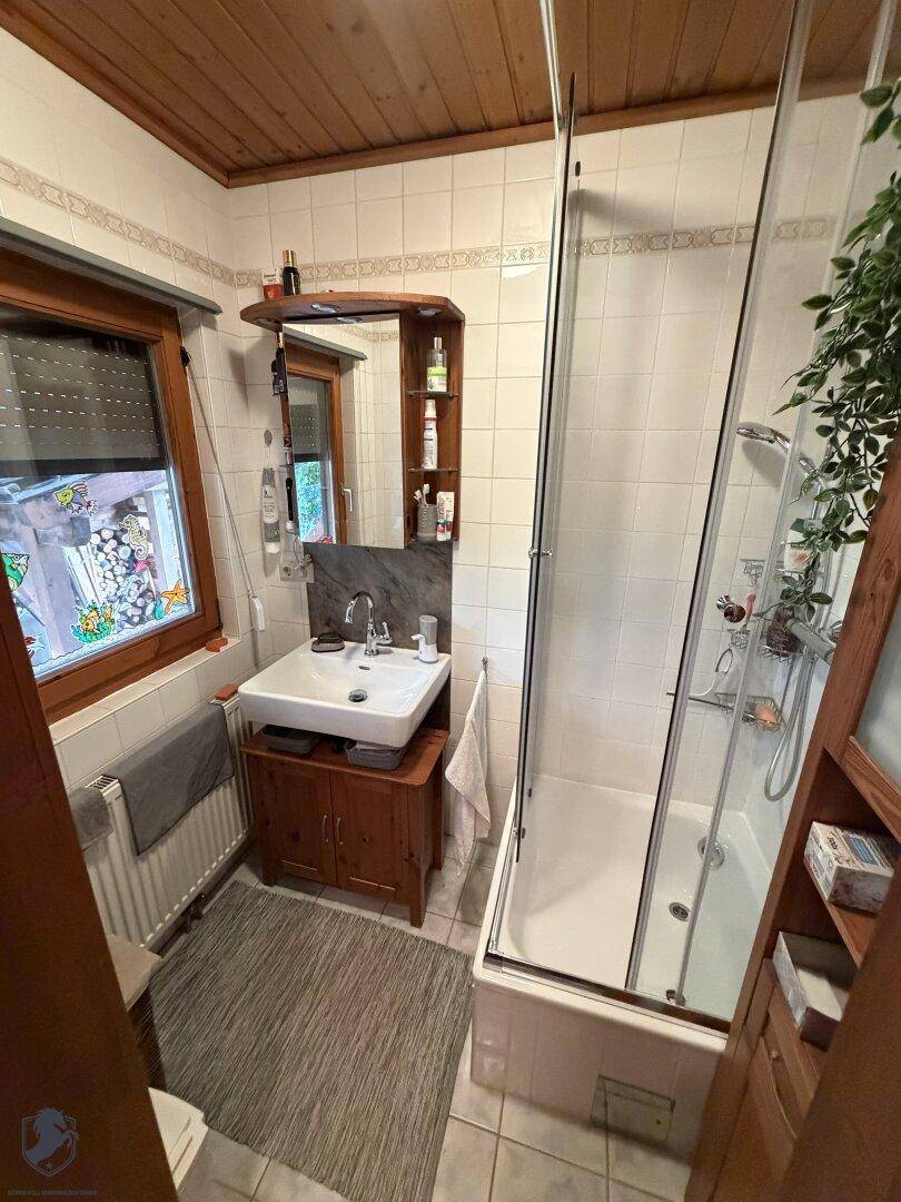 Badezimmer mit Dusche in Badewanne, Waschbecken und Fenster, ausgestattet mit Fliesen.