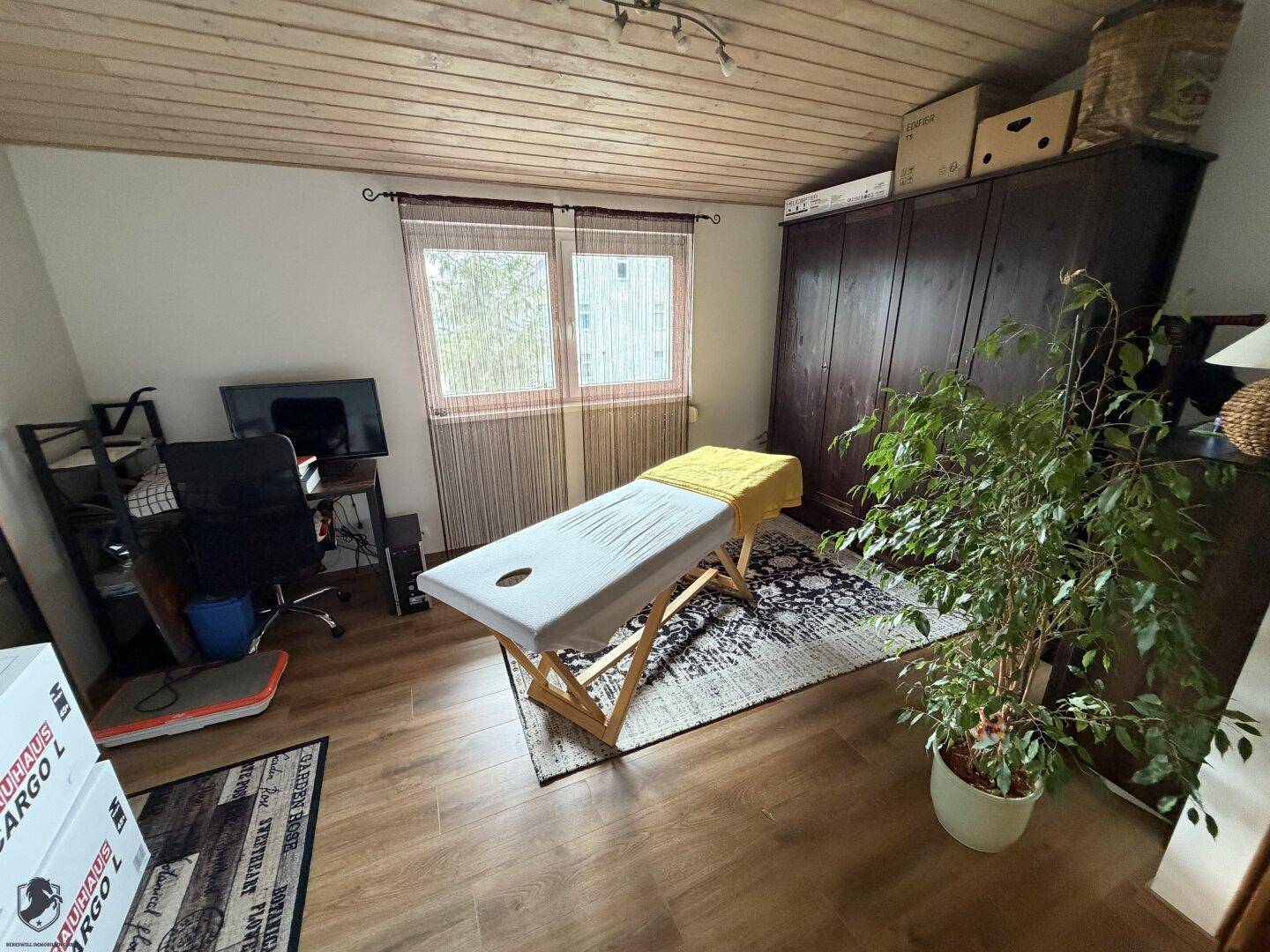 Vielseitiger Raum mit Massageliege, Schreibtisch und großem Kleiderschrank, unter einer Holzdecke.