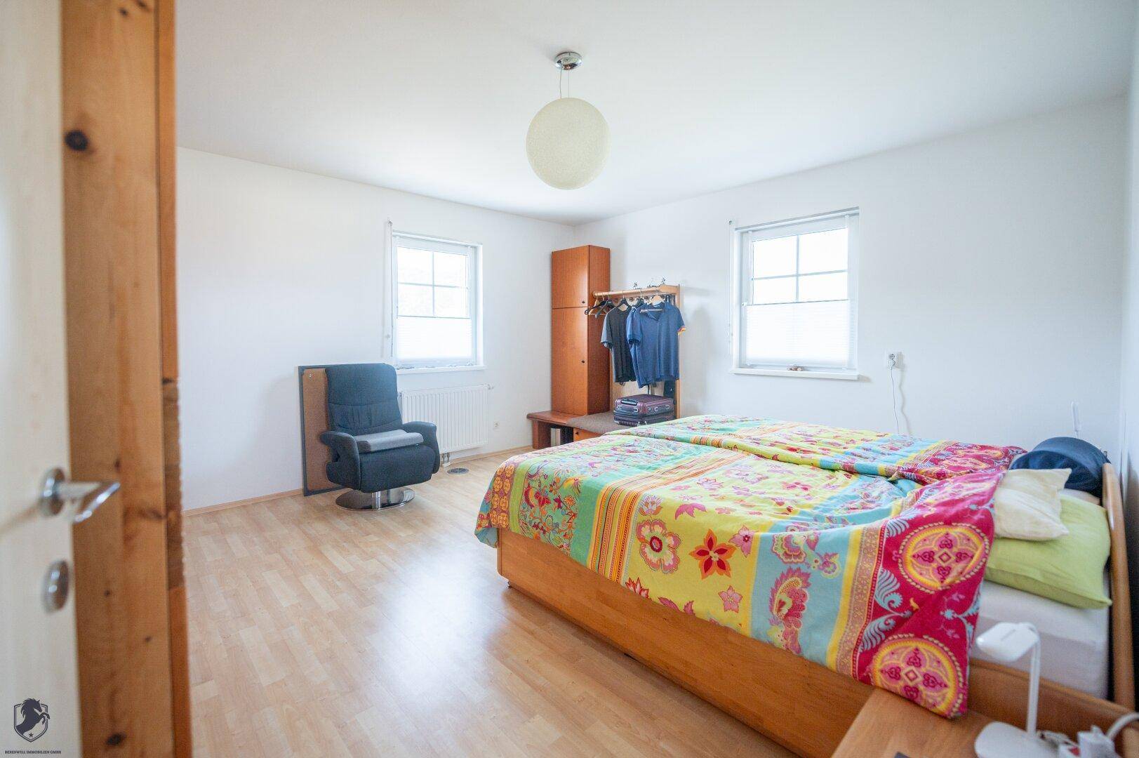 Schlafzimmer mit Doppelbett, bunten Decken und hellem Laminatboden, gut beleuchtet durch Fenster.