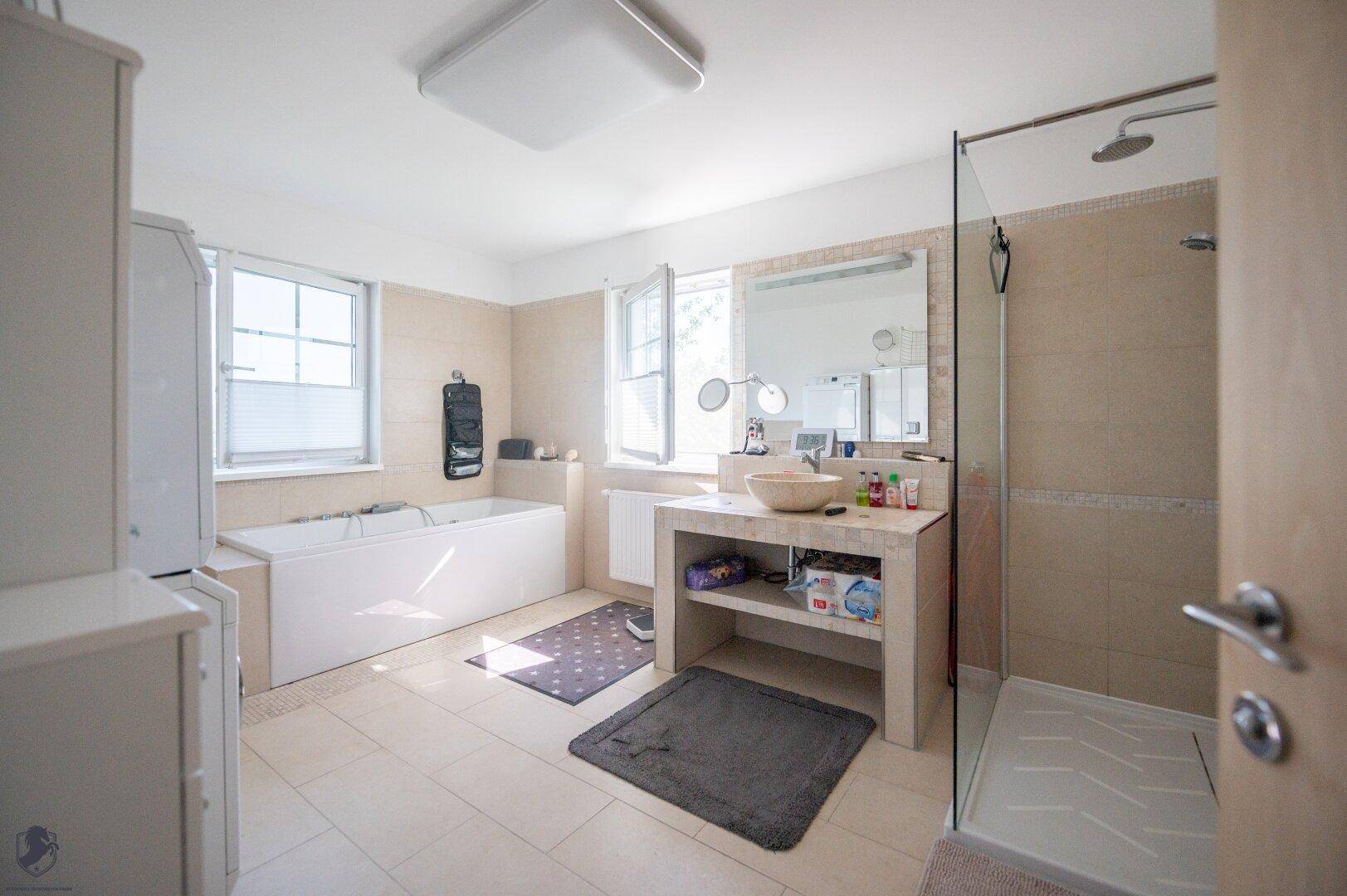 Geräumiges Badezimmer mit Badewanne, Dusche und hellem Fliesenboden, gut beleuchtet durch Fenster.