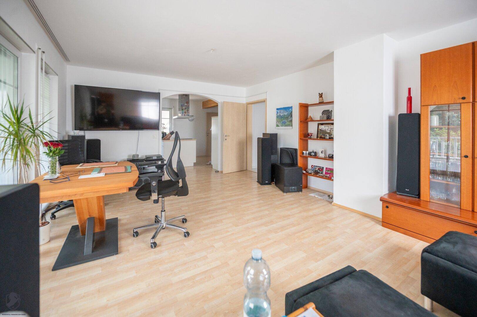 Wohnzimmer mit großem Fernseher, Arbeitsbereich und offenem Übergang zu weiteren Räumen.