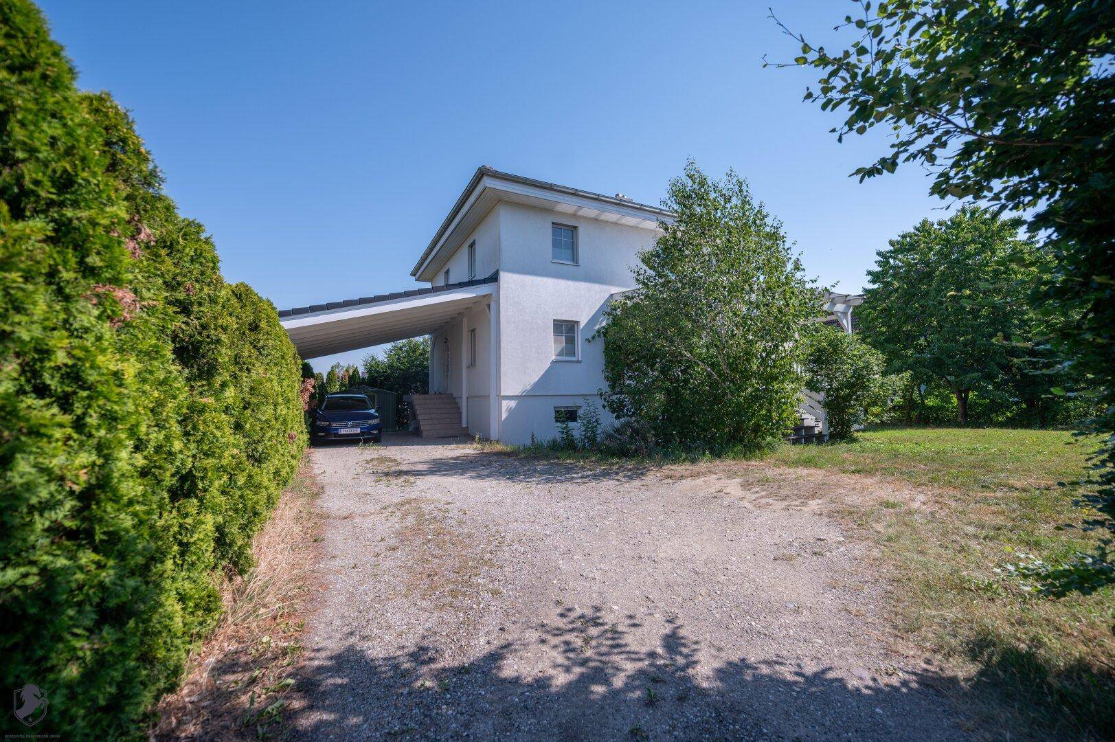 Außenansicht des Hauses mit Carport, Kiesauffahrt und umgebender grüner Hecke.