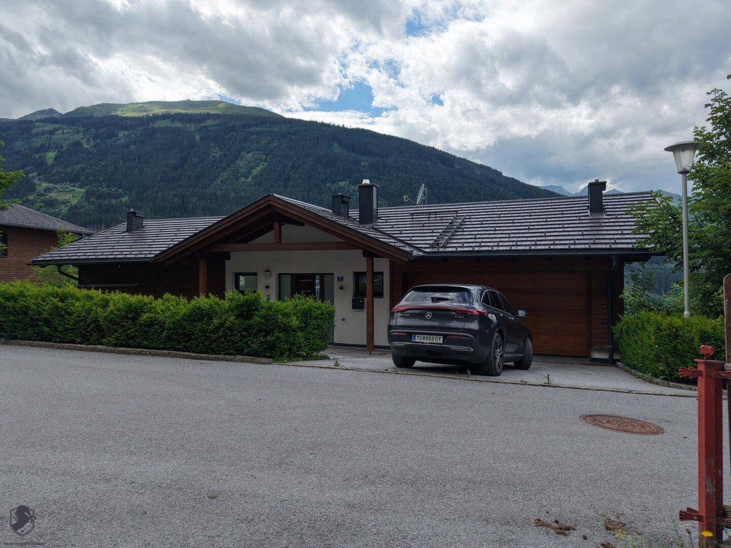 Vorderansicht eines modernen Hauses mit Holzelementen, dunklem Dach und gepflegter Hecke vor Bergkulisse.