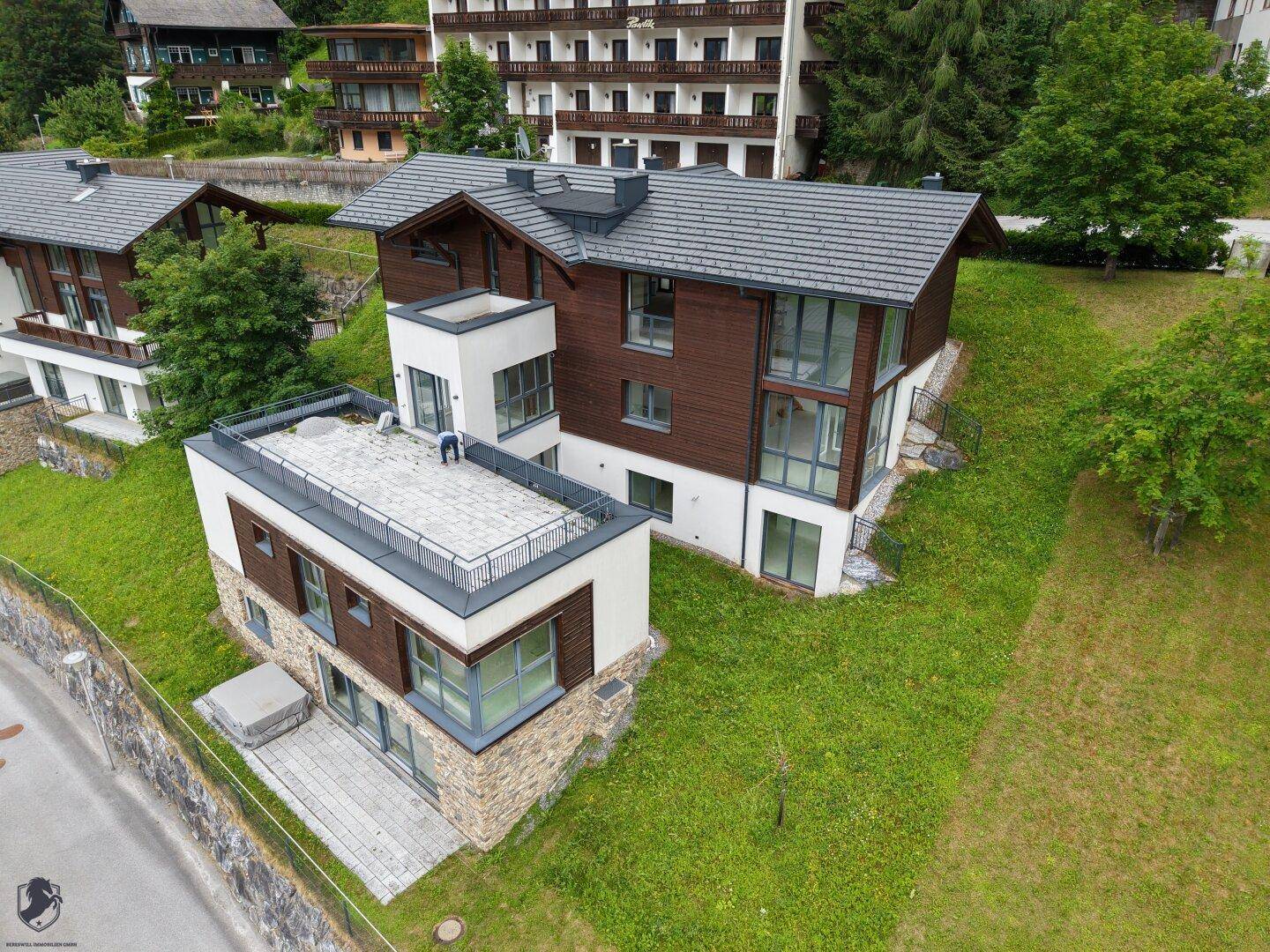 Luftaufnahme von modernen Häusern mit dunklen Dächern und großen Fensterfronten, eingebettet in grüne Hügel.
