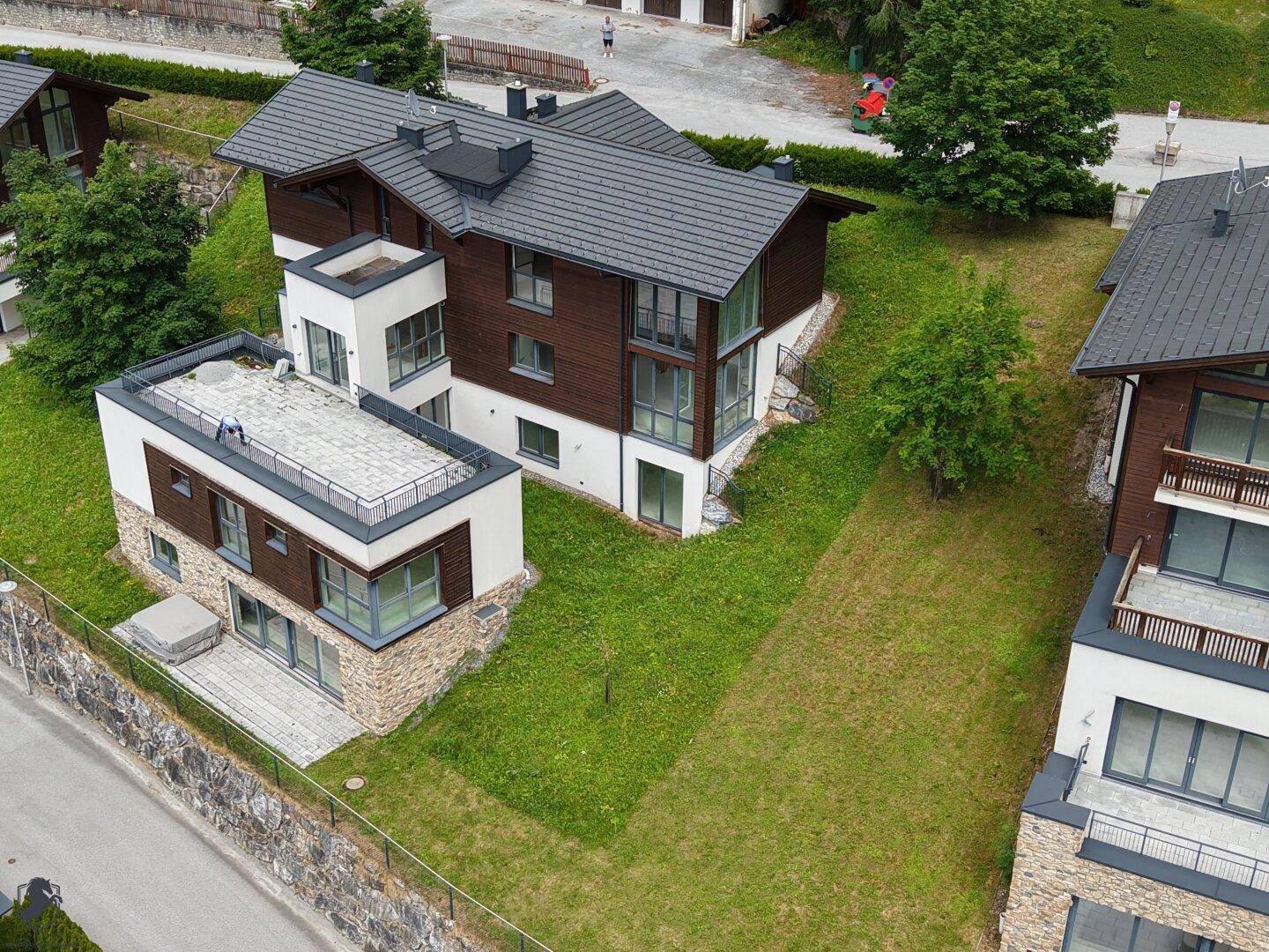 Luftaufnahme von modernen Häusern mit dunklen Dächern und großen Fensterfronten, umgeben von grünen Hängen.