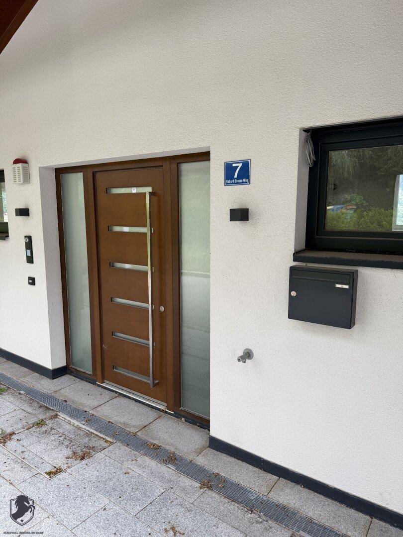 Moderner Hauseingang mit Holztür, seitlichen Glaspaneelen und einer Hausnummer an der weißen Fassade.