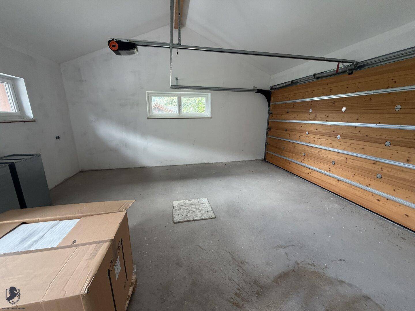 Geräumige, leere Garage mit Betonboden, Holz-Garagentor und zwei Fenstern für Tageslicht.