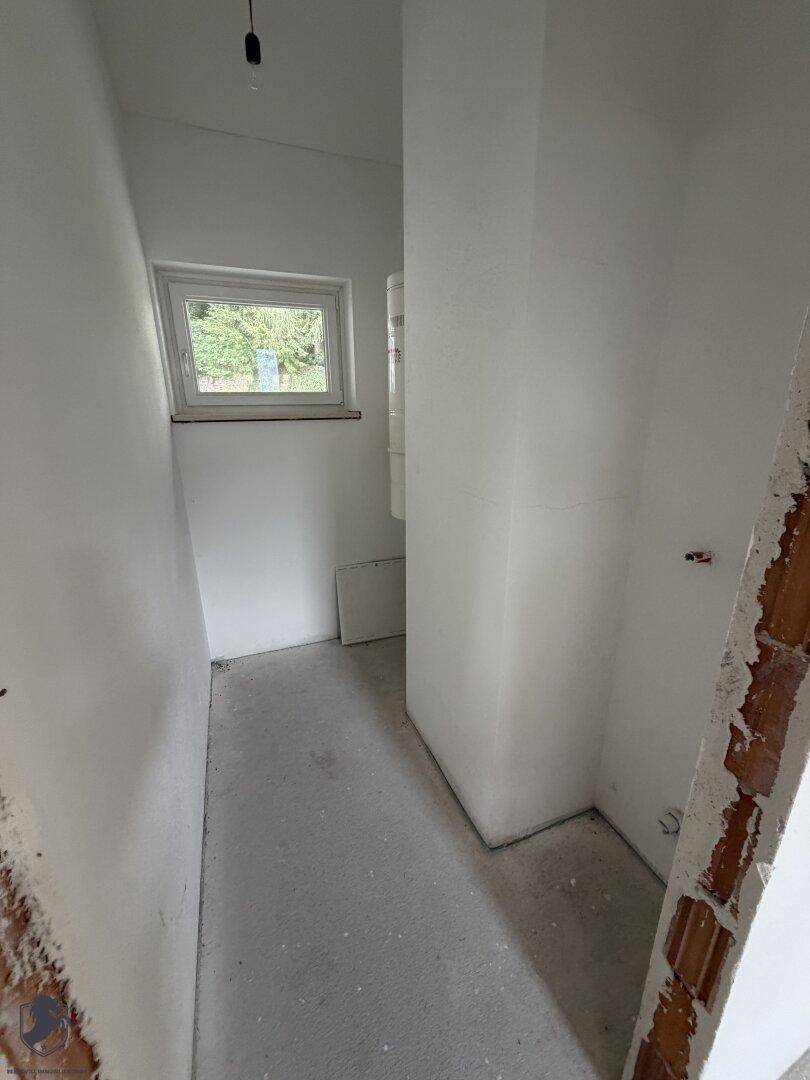 Schmaler Hauswirtschaftsraum mit weiß gestrichenen Wänden, Betonboden und einem kleinen Fenster.