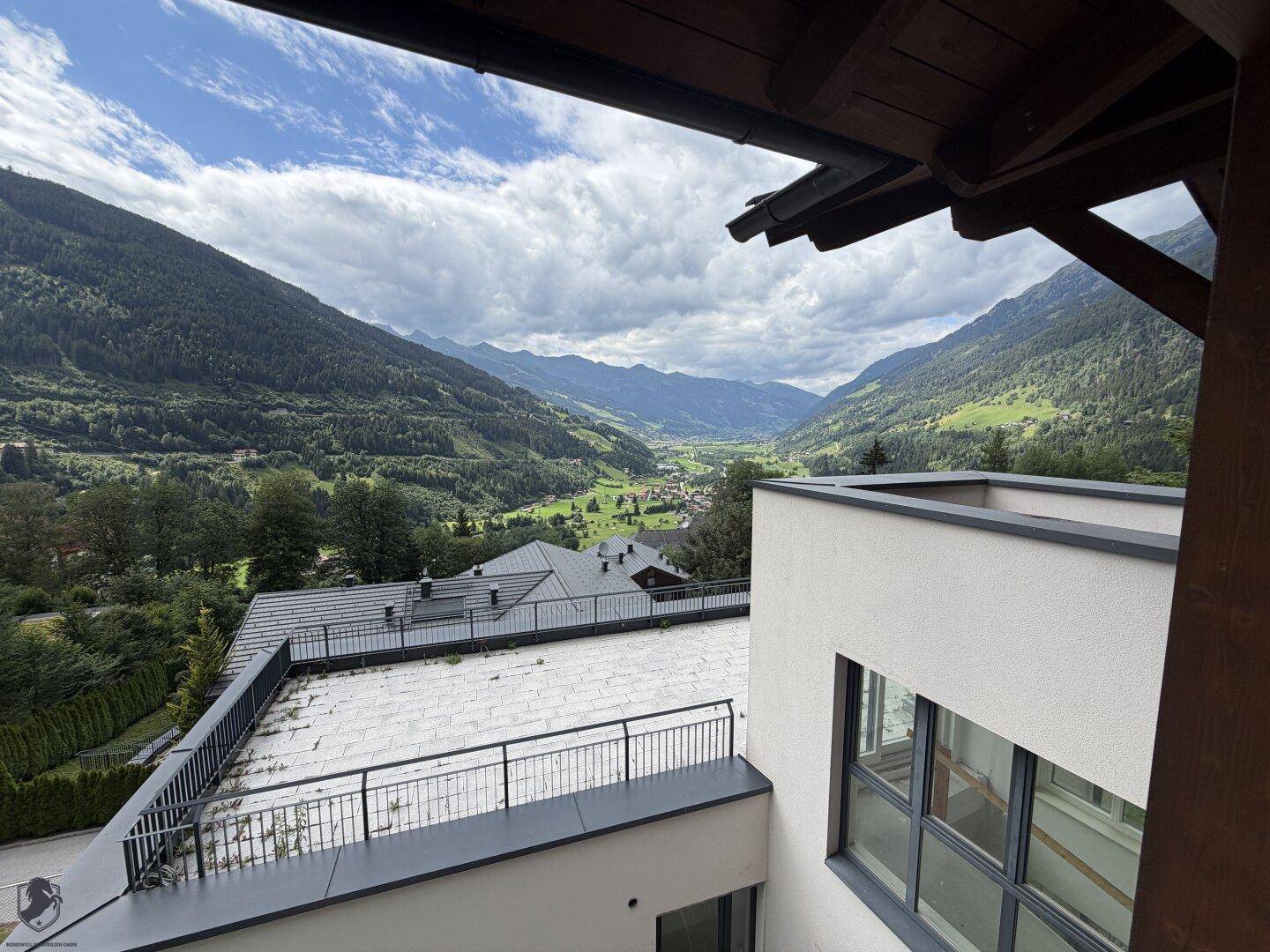 Weitläufiger Blick von einer Terrasse auf ein grünes Tal und die umliegende Berglandschaft unter bewölktem Himmel.