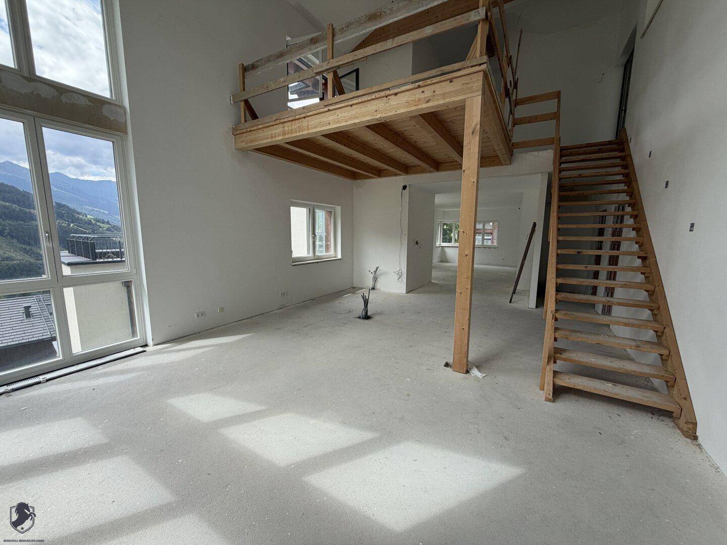 Geräumiger, heller Wohnbereich im Rohbau mit hohen Decken, Betonboden und großen Fenstern mit Bergblick.