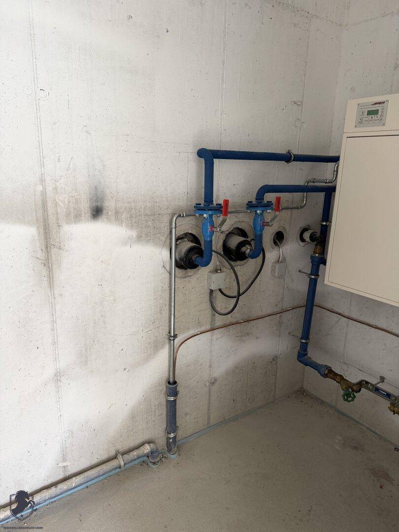 Technikraum im Keller mit sichtbaren blauen Rohren und einem Heizungs- oder Steuerungssystem an der Wand.