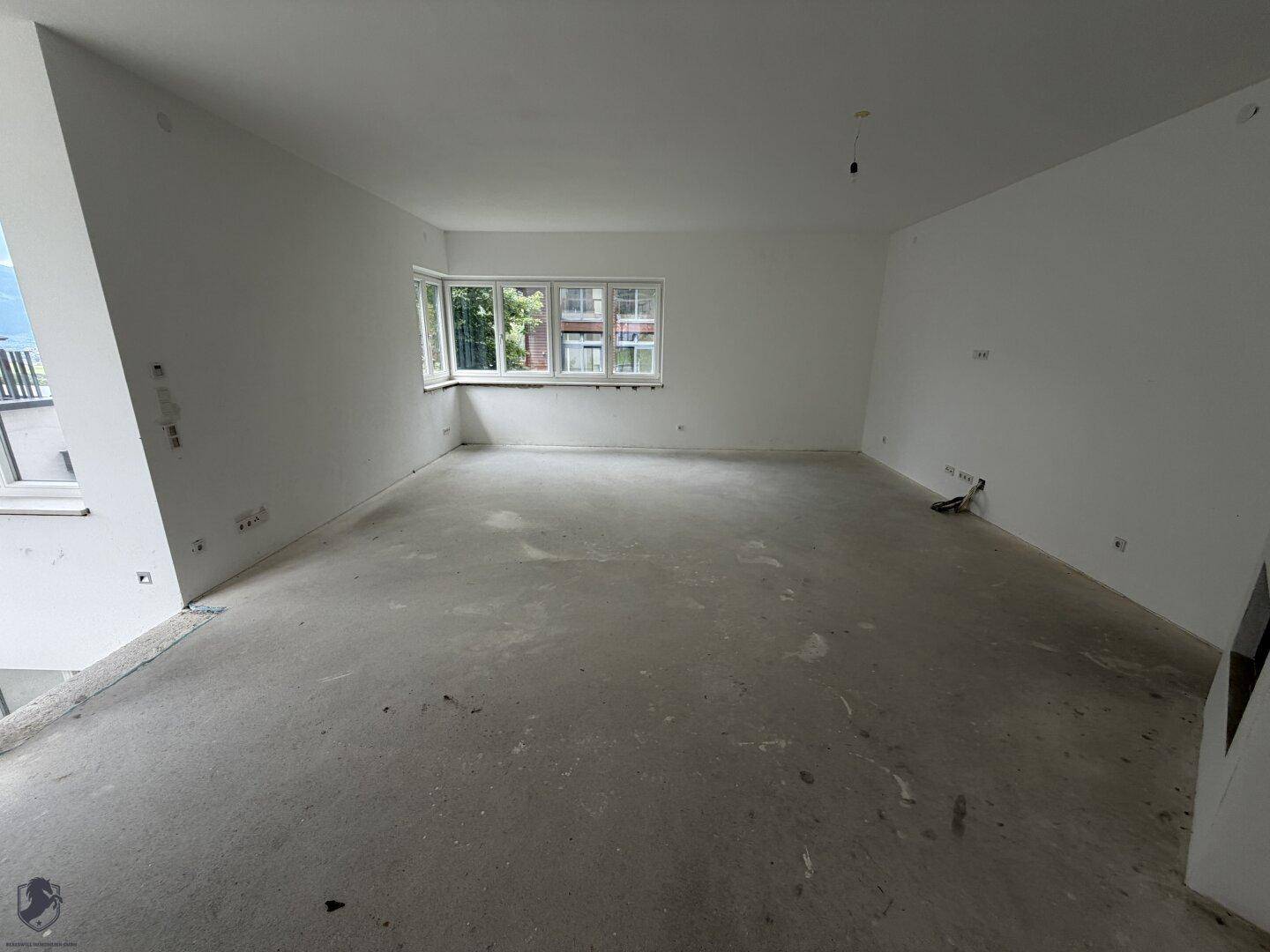 Großer, heller Wohnbereich im Rohbau mit Betonboden und einer Fensterfront, die viel Tageslicht hereinlässt.