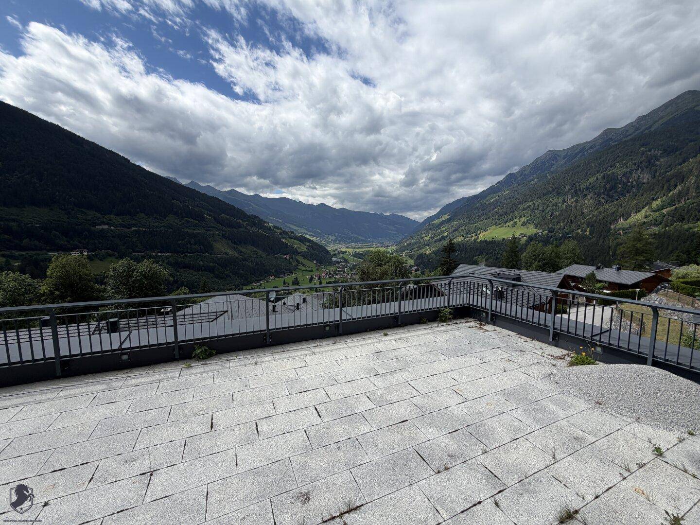 Große, gepflasterte Terrasse mit Metallgeländer und weitem Blick auf ein grünes Tal und die Berge.