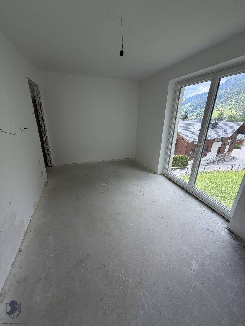 Heller Raum im Rohbau mit Betonboden und einer großen Fensterfront, die viel Tageslicht und Bergblick bietet.