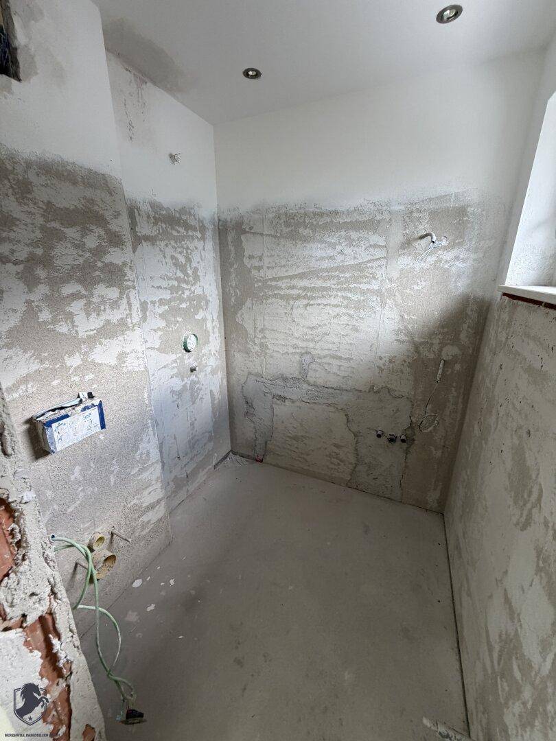 Badezimmer im Rohbau mit unverputzten Wänden, Betonboden und vorbereiteten Anschlüssen für Sanitäranlagen.