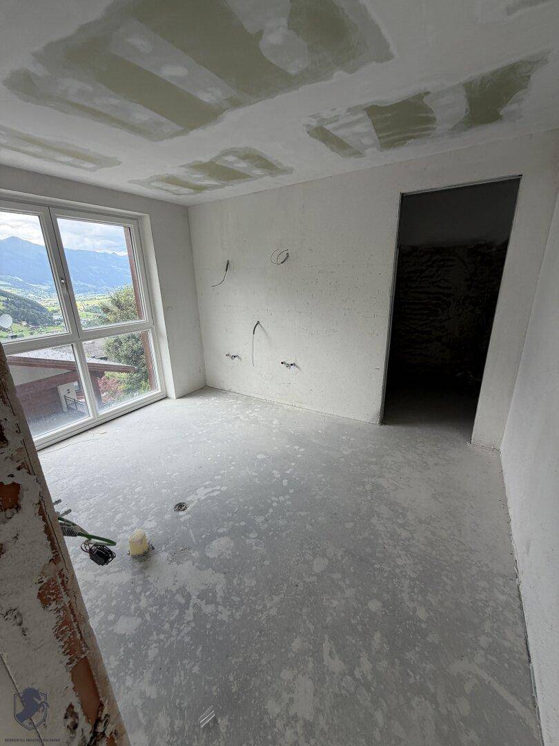 Unfertiger Raum mit großem Fenster, Betonboden und Blick auf die bergige Landschaft.