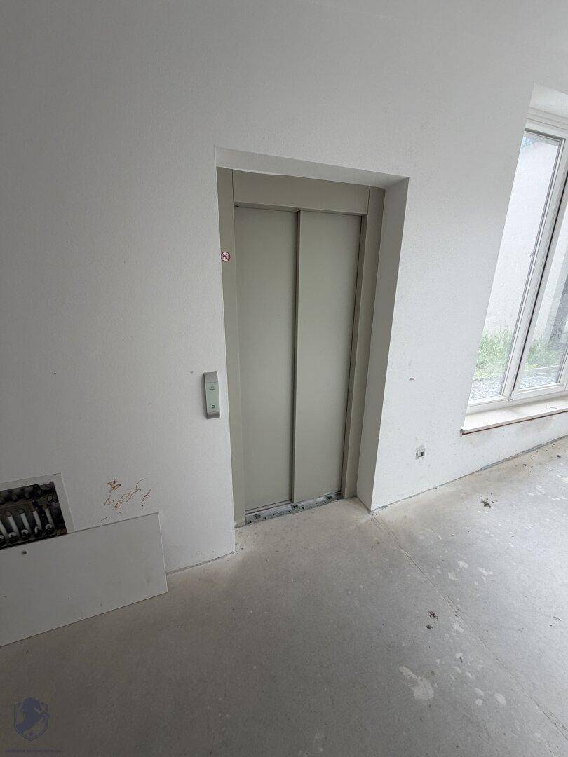 Aufzugstür in einem unfertigen Flur mit weiß gestrichenen Wänden und Betonboden.