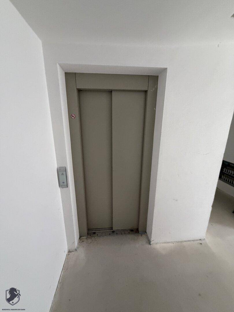 Aufzugstür in einem unfertigen Flur mit weiß gestrichenen Wänden und Betonboden.