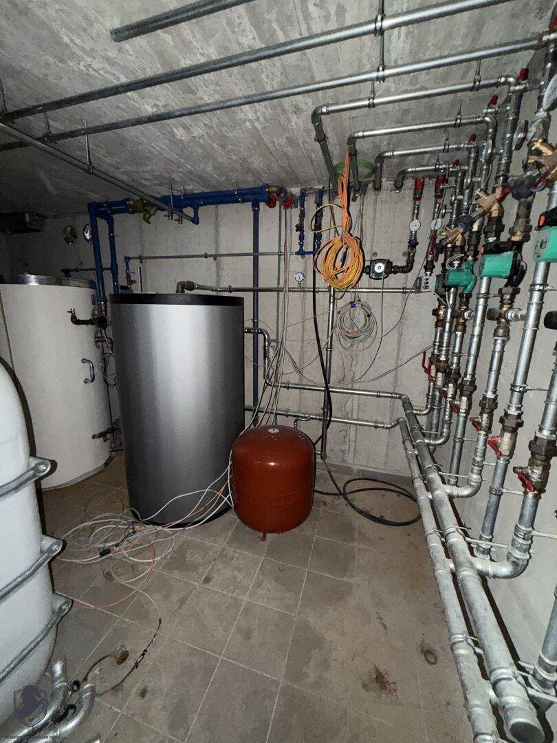 Technikraum im Keller mit zahlreichen Rohren und Warmwasserspeichern auf gefliestem Boden.