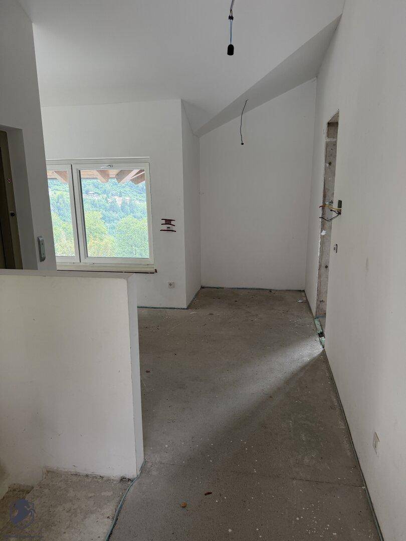 Unfertiger Innenraum mit Betonboden, weißen Wänden und Fensterblick ins Grüne, noch im Rohbau.