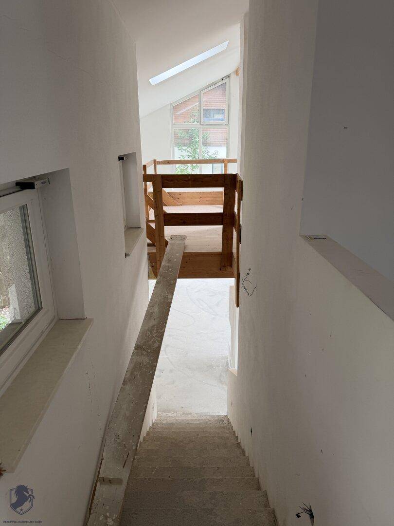 Unfertiges Treppenhaus mit Betonstufen und Blick auf eine obere Etage mit Holzgeländer und Fenster.