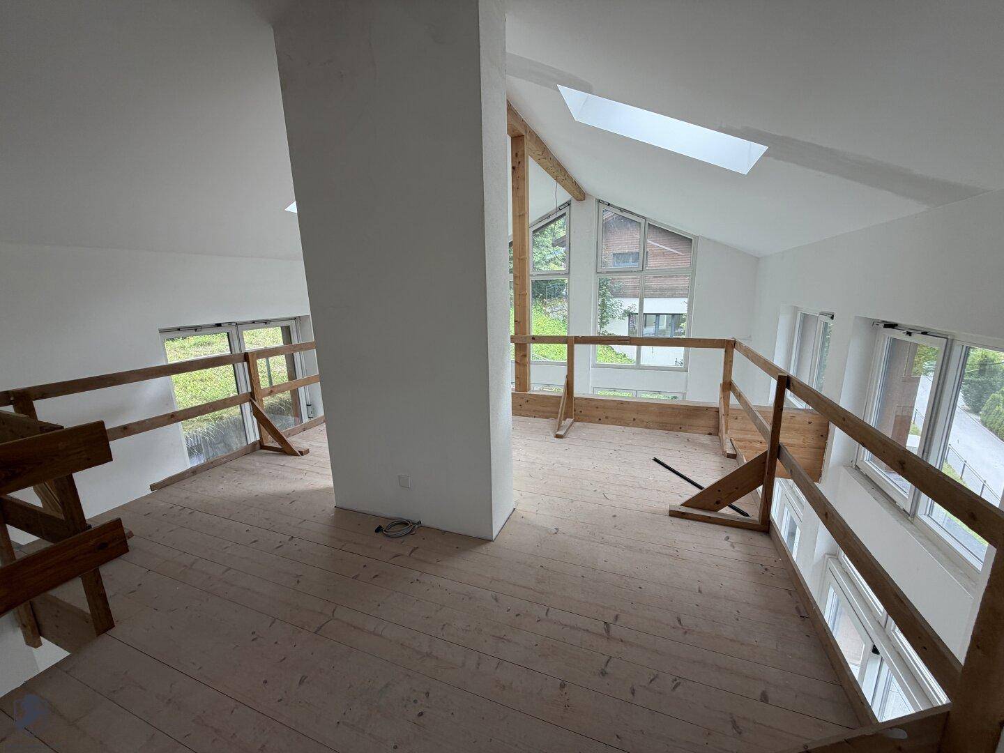 Geräumiger, unfertiger Wohnbereich mit hohen Decken, Holzboden und großen Fenstern, lichtdurchflutet.