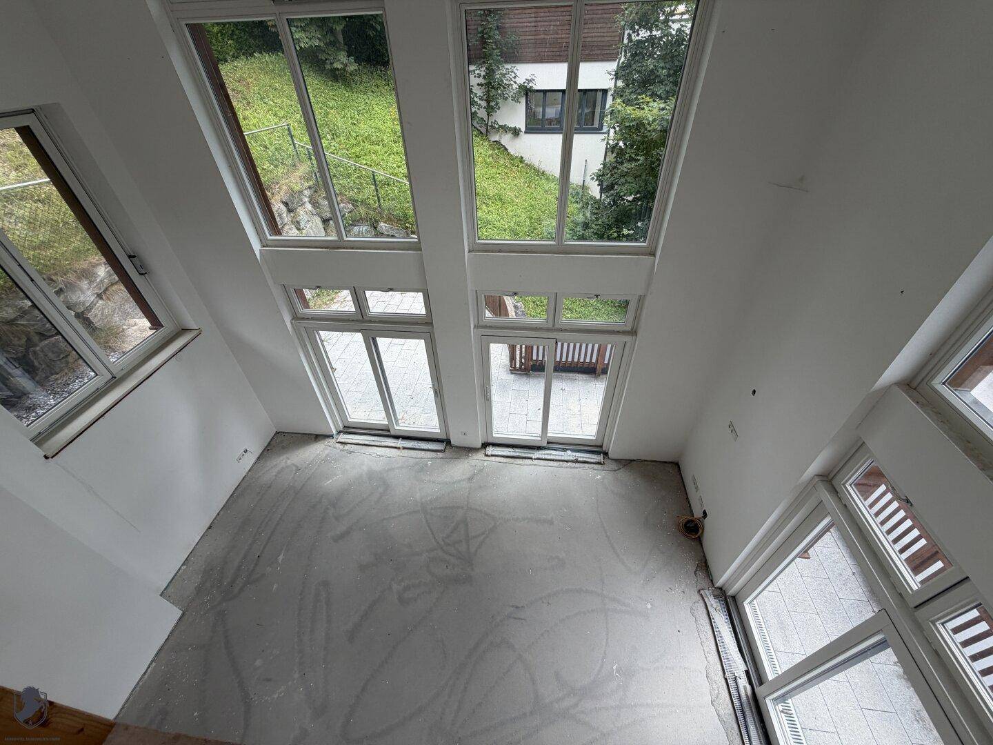Heller Wohnbereich im Rohbau mit hohen Fenstern und Blick auf die grüne Umgebung, viel Tageslicht.