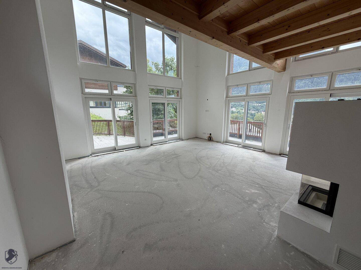 Geräumiger Wohnbereich im Rohbau mit hohen Decken, großen Fenstern und modernem Kamin.