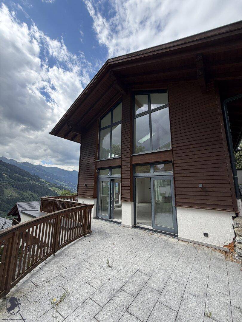 Terrasse vor einem modernen Holzhaus mit großen Fenstern und Bergblick, viel Tageslicht.