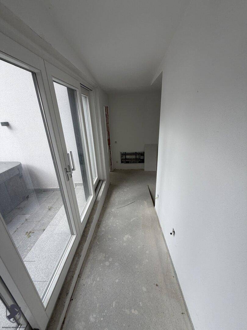 Langer Flur im Rohbau mit Betonboden und großen Fenstern, die auf eine Terrasse blicken.