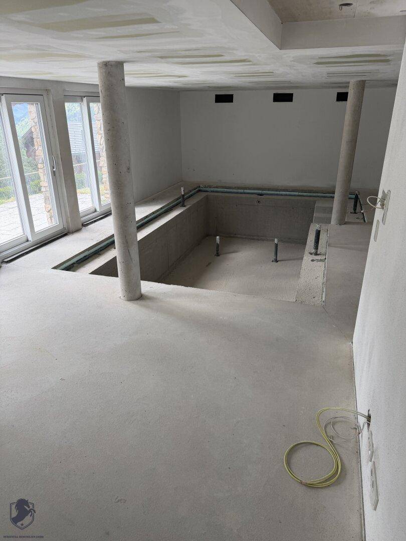 Großer Raum im Rohbau mit vorbereitetem Poolbereich, Säulen und großen Fenstern.