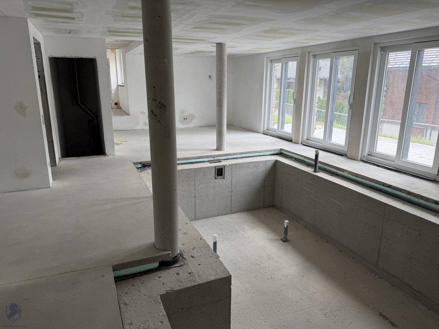 Geräumiger Rohbau mit einem zukünftigen Pool, tragenden Säulen und viel Tageslicht durch große Fenster.
