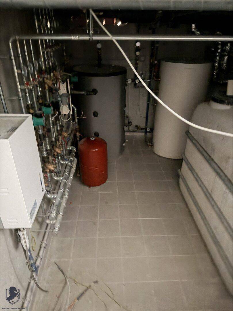 Technikraum mit Heizungsanlage, Warmwasserspeichern und zahlreichen Rohrleitungen auf Fliesenboden.