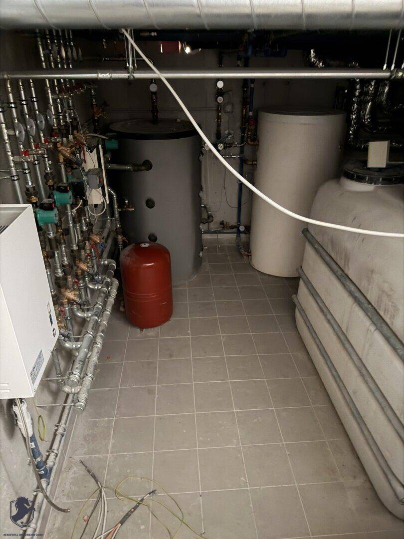 Heizungsraum mit modernen Anlagen, großen Tanks und komplexen Rohrleitungssystemen auf Fliesen.