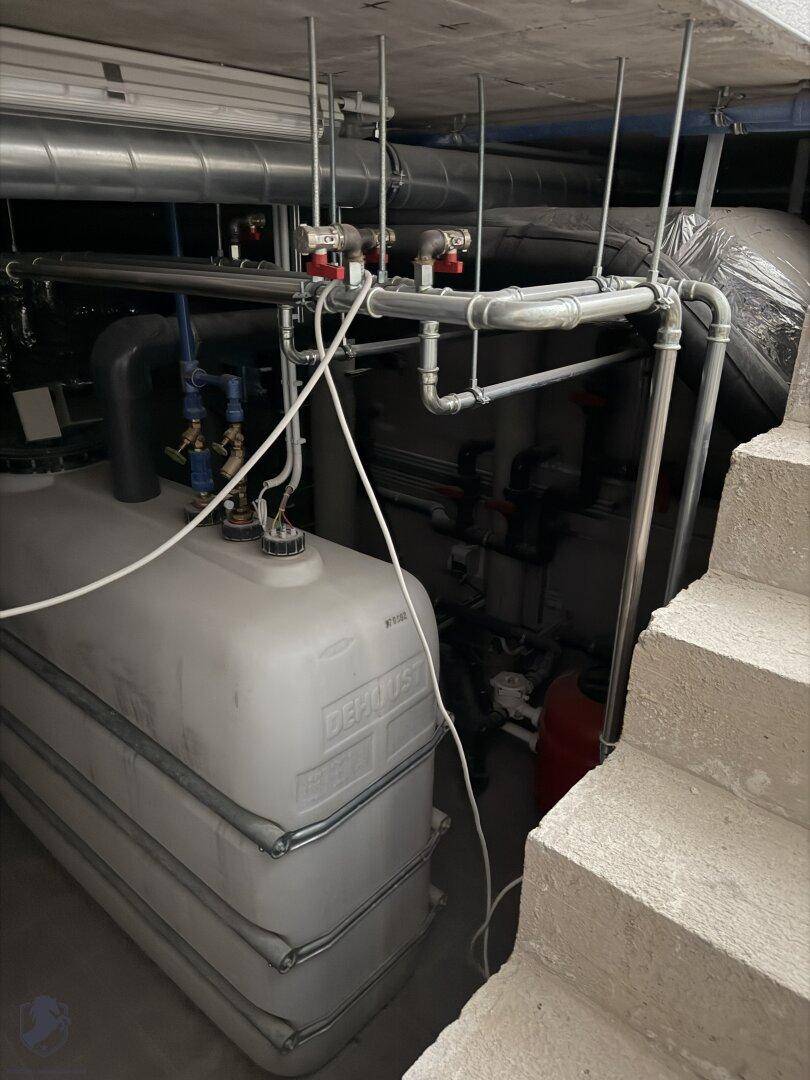 Kellerraum mit großen Wassertanks und Rohrleitungen, daneben eine Betontreppe.
