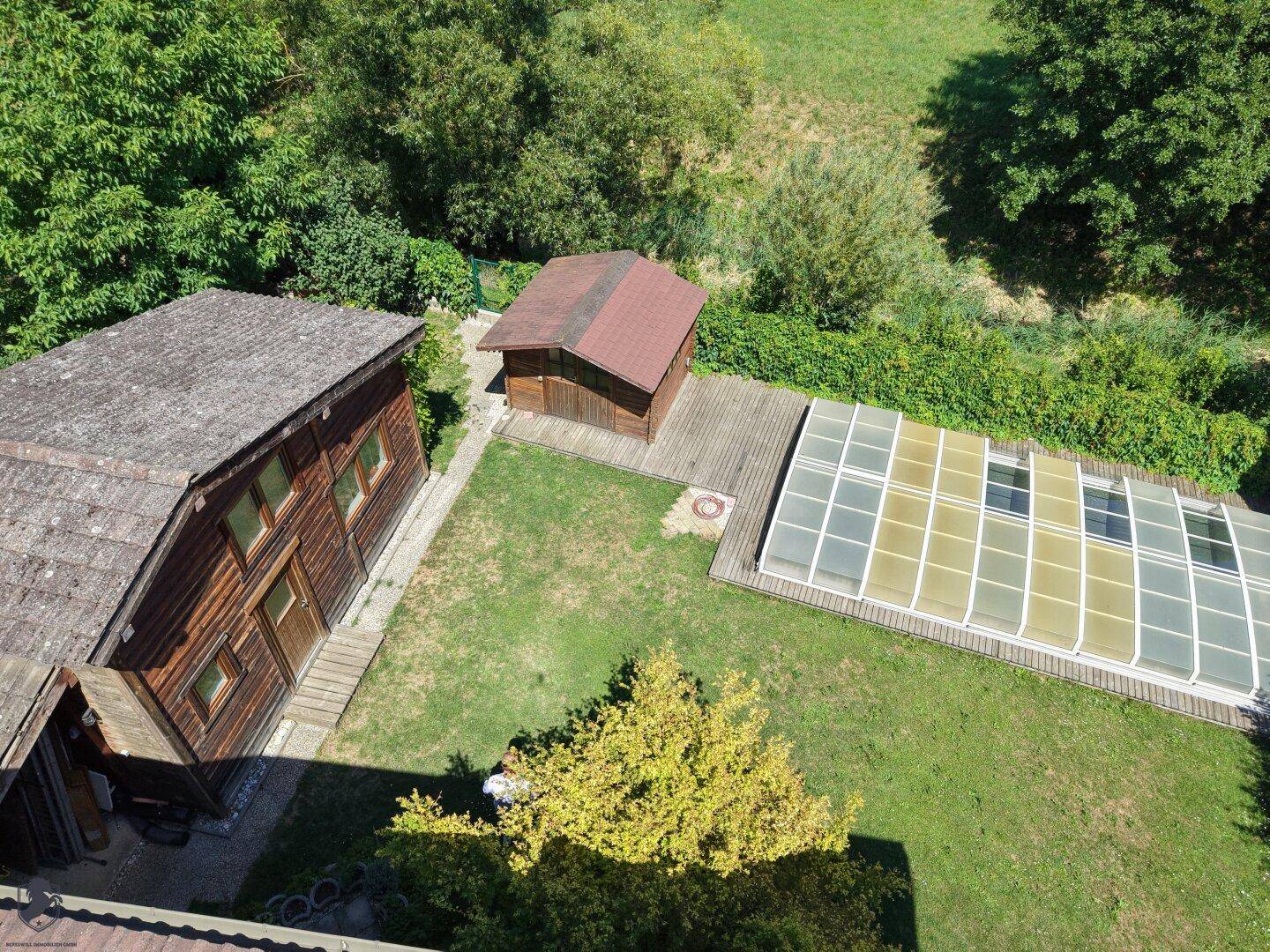 Blick auf das Holzhaus, ein Nebengebäude, den überdachten Pool und die angrenzende Holzterrasse.
