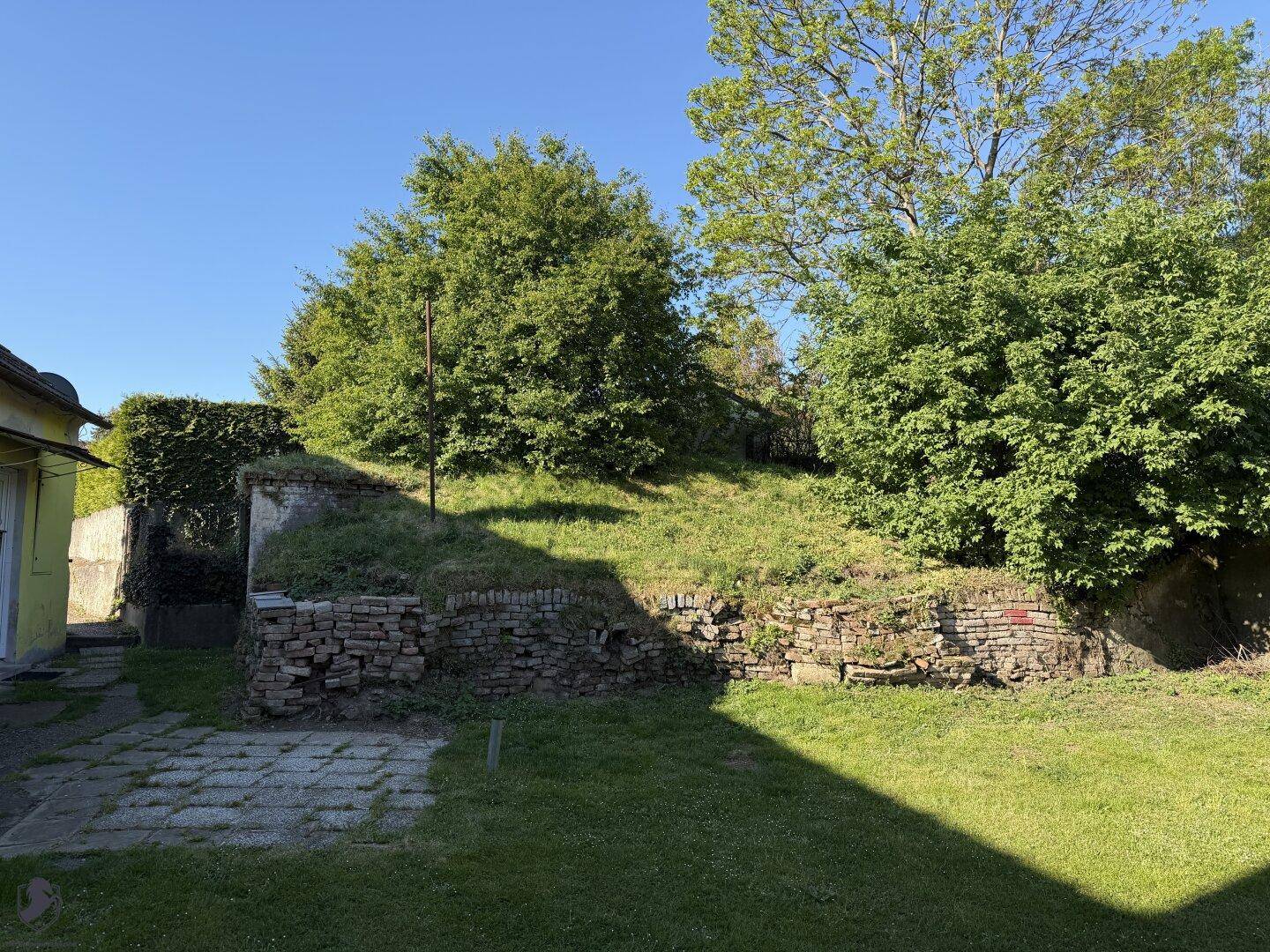 Grüner Gartenbereich mit alten Steinmauern und üppiger Vegetation unter blauem Himmel.